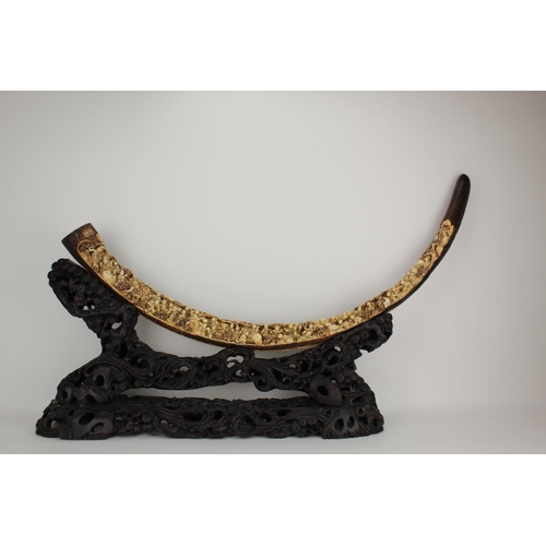 A large carved woolly mammoth tusk of curved form with rich brown and tan hues, finely carved in the traditional Chinese manor with a panoply of musicians, tradesman, trees and buildings, signed to the end and engraved to the reverse with a continuous scene of children playing draughts, figures working, tigers, trees and fences, text and signature, 127cm long, approx 100cm tip to end, 7.5cm dia at base, on a carved wooden stand with gnarled trees and scrolls