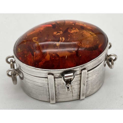 A 925 silver trinket pot set with large amber cabochon to lid. Silver hinge lidded pot in the form of an oval shaped banded casket with drop down handles and drop over latch. Fully hallmarked to underside with London assay mark and G Ltd makers mark. Both base and lid stamped 925 and SP. Approx. 3cm tall x 5cm long. Total weight approx. 45.4g.